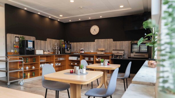 A modern hotel lobby with wooden furniture, a clock on the wall, and a coffee machine. - Plaza Inn Hannover City Nord (Hannover)