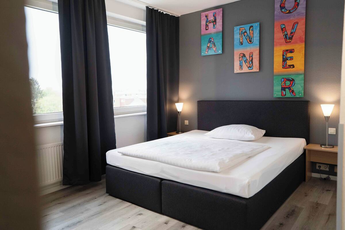 A neatly made bed in a modern room with a large window, dark curtains, and wall art spelling out "HAPPY HOME."