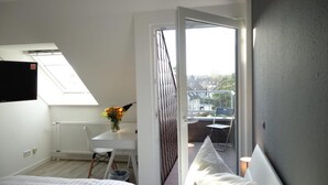 A bedroom with a skylight, a white desk with a vase of flowers, a bed, and a view of a balcony with a chair. - Plaza Inn Hannover City Nord (Hannover)