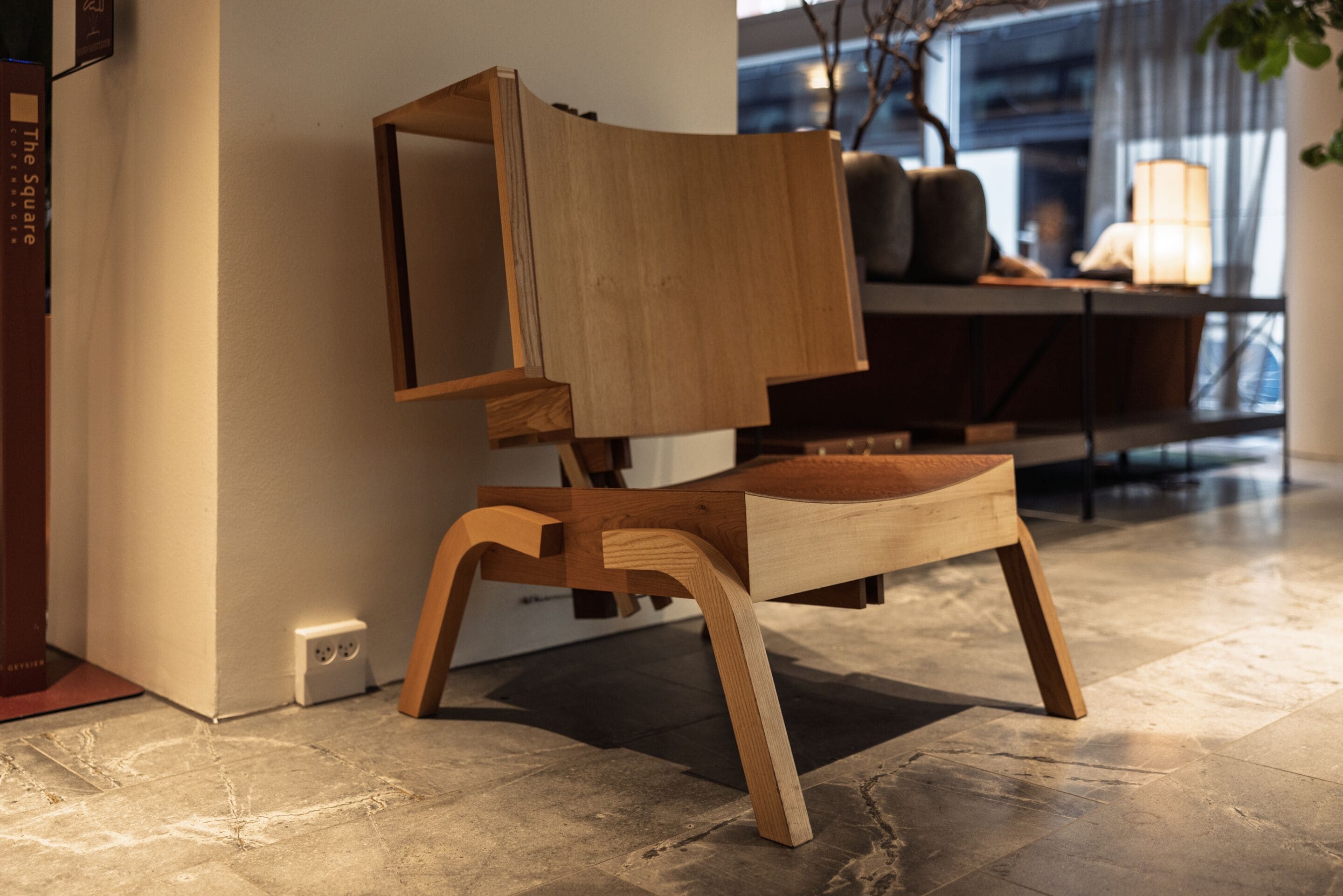 A modern wooden chair with a unique design, placed in a well-lit room with a tiled floor and a window in the background.