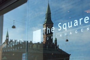 A window with the text "The Square Copenhagen" and a reflection of a building with a spire.