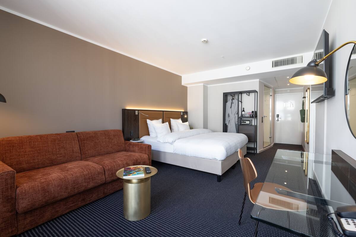 A hotel room with a bed, a sofa, a glass table, and a chair.