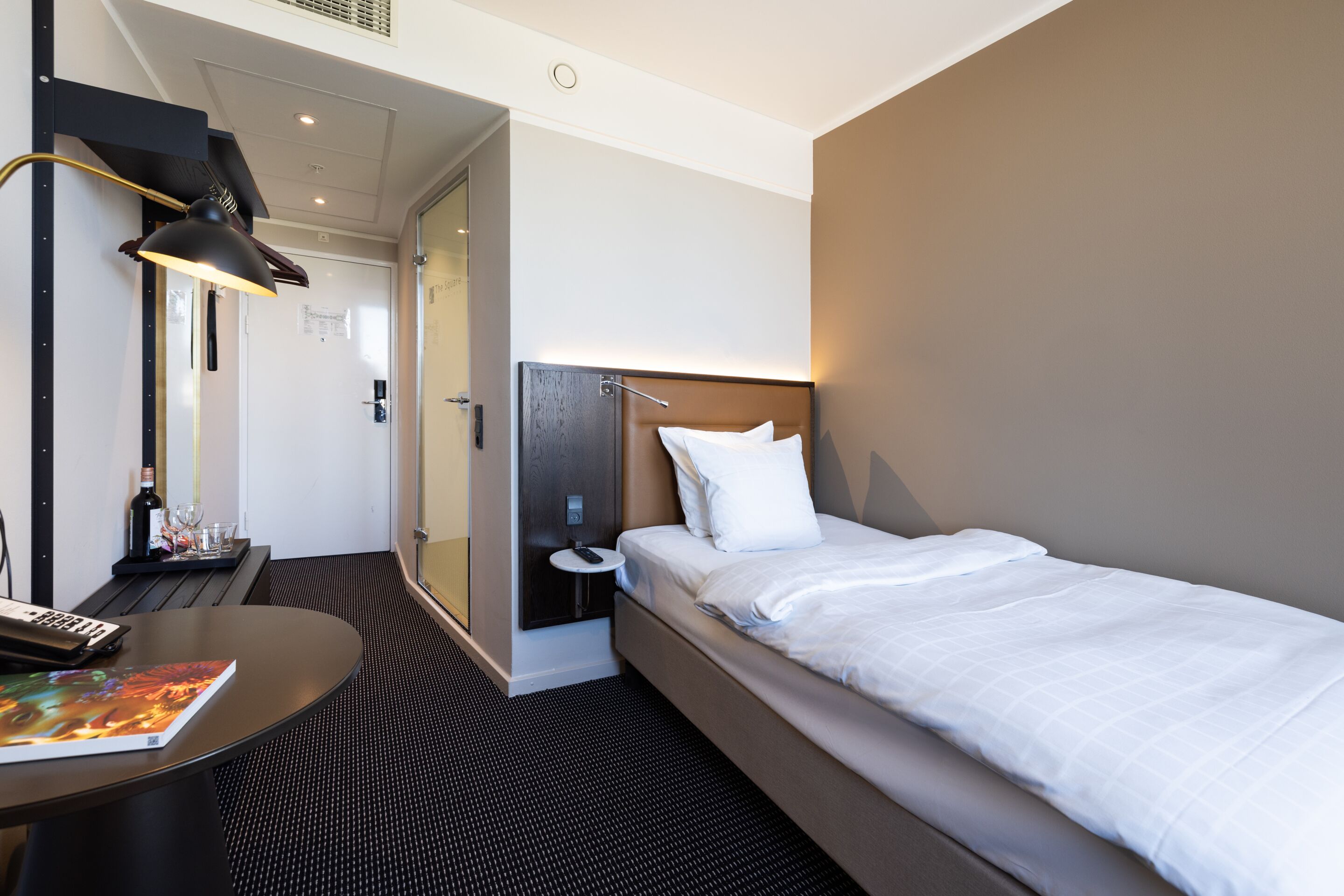 A modern hotel room with a single bed, a desk with a computer and a book, a small table with a bottle and glasses, and a wall-mounted lamp.