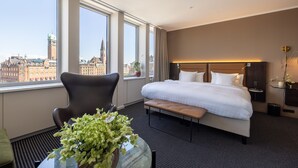 A hotel room with a large bed, a chair, a small table with a plant, and a view of a cityscape through large windows. - The Square (Copenhagen)