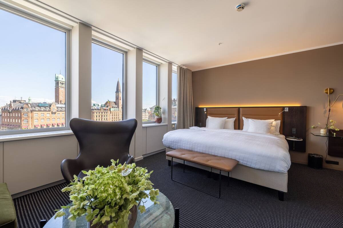 A hotel room with a large bed, a chair, a small table with a plant, and a view of a cityscape through large windows.