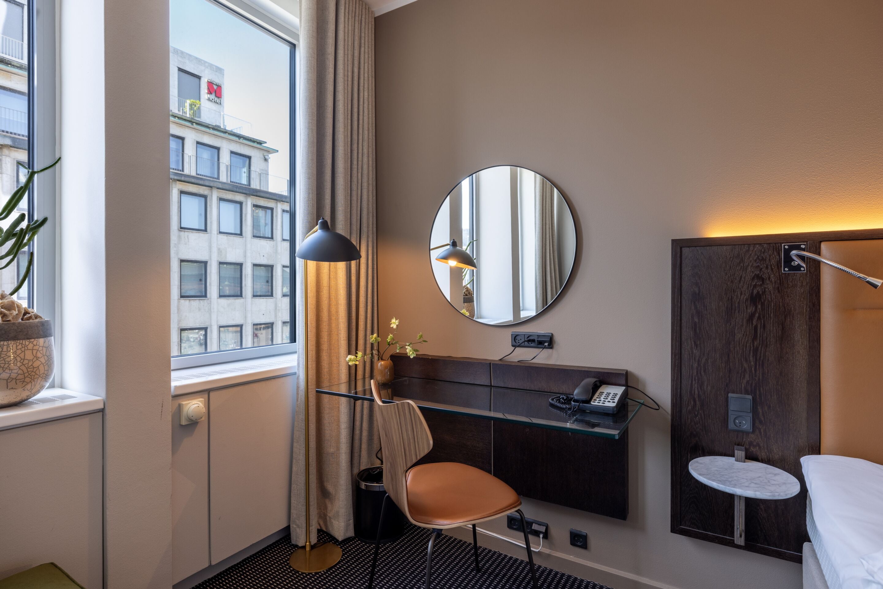 Ein Hotelzimmer mit einem Bett, einem runden Spiegel, einem Schreibtisch mit Telefon, einem kleinen runden Tisch und einem Fenster mit Vorhängen.
