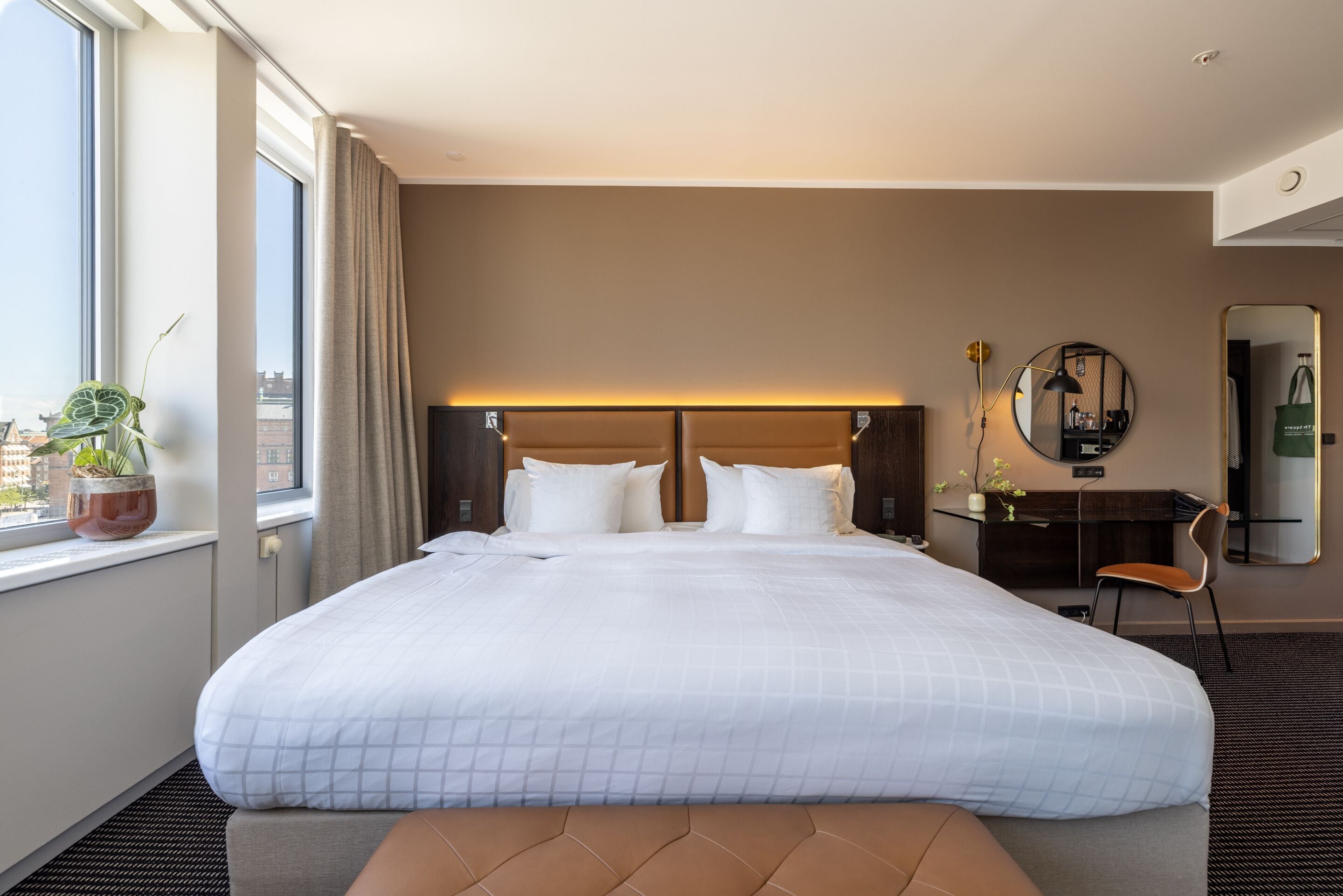 A hotel room with a large bed, a desk, a chair, a mirror, and a window with a view of buildings.