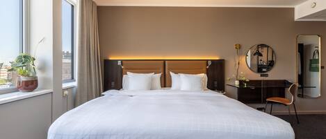 A hotel room with a large bed, a desk, a chair, a mirror, and a window with a view of buildings.