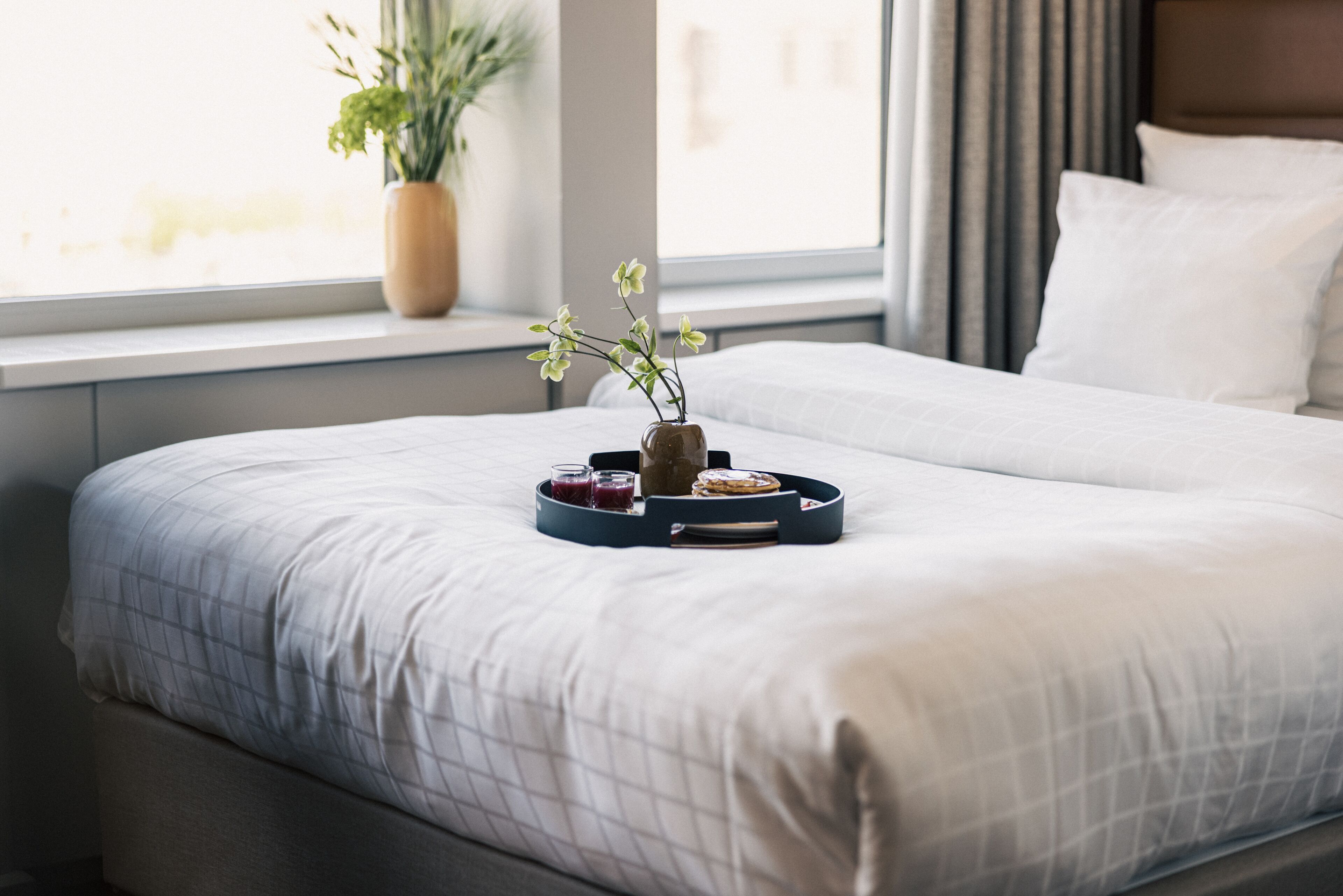 A neatly made bed with a tray of breakfast items, a vase with flowers, and a window with a plant.