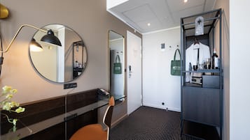 Una habitación de hotel con un espejo redondo, un estante de vidrio, una silla, una mesita, un perchero colgante y un armario empotrado.