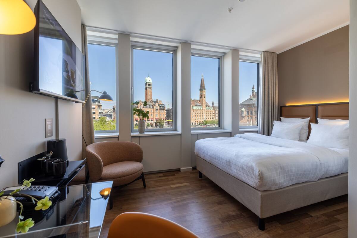 A hotel room with a large bed, a desk with a computer, a chair, and large windows offering a city view.