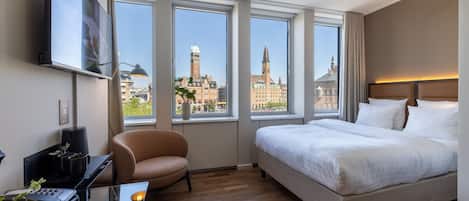 A hotel room with a large bed, a desk with a computer, a chair, and large windows offering a city view.