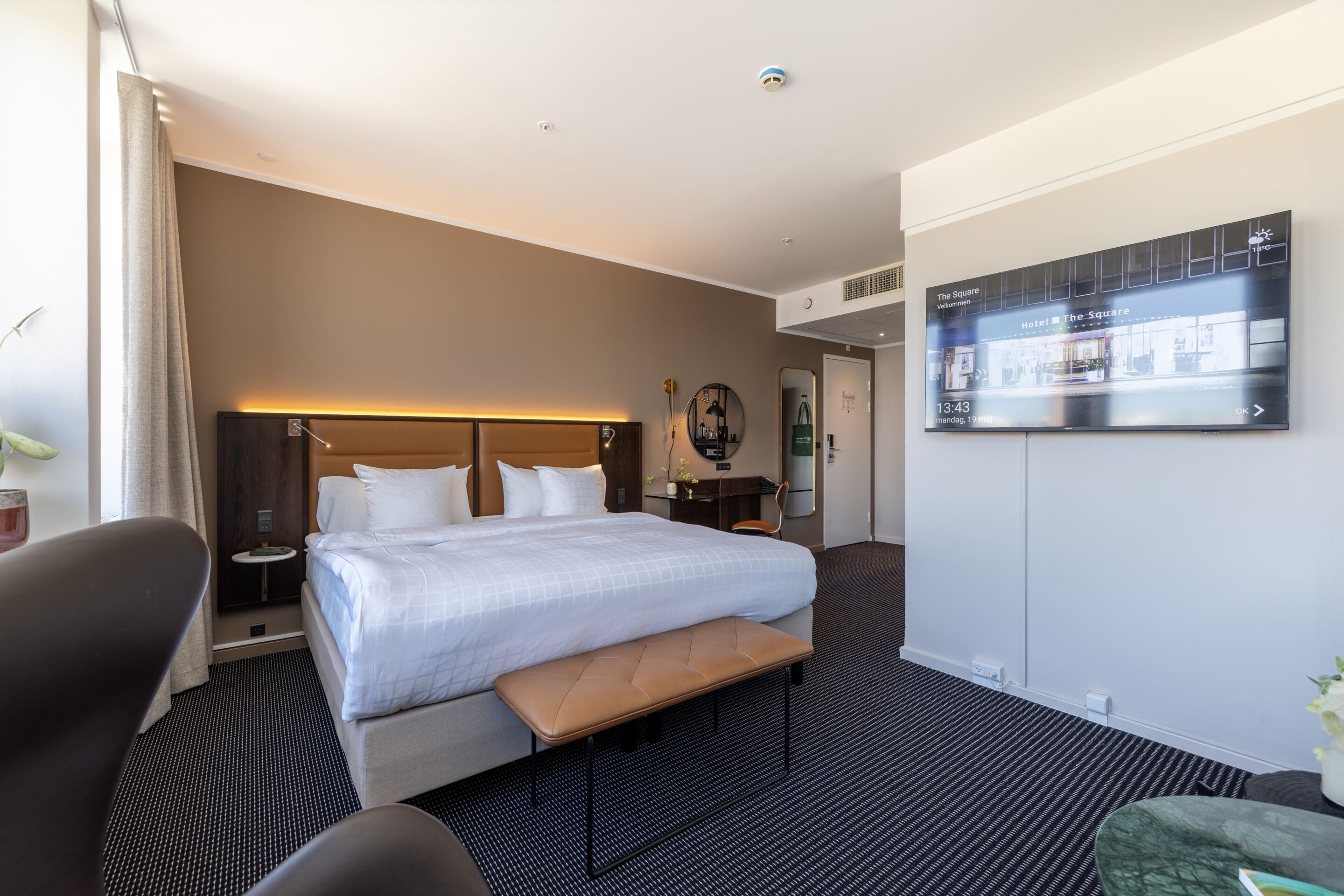 Ein Hotelzimmer mit Bett, Sitzbank, Schreibtisch, Stuhl, Fernseher und einer Pflanze.