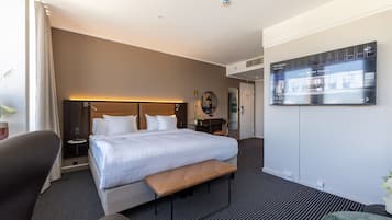 Ein Hotelzimmer mit Bett, Sitzbank, Schreibtisch, Stuhl, Fernseher und einer Pflanze.