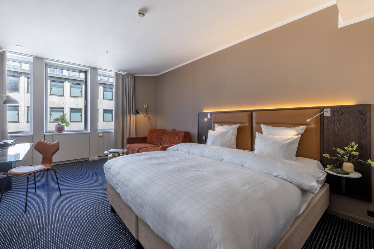 A hotel room with a large bed, a desk with a chair, a small table, and a potted plant.