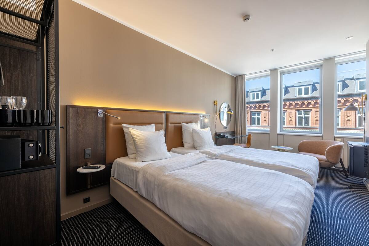 A hotel room with two beds, a minibar, a round table, a chair, and large windows with a view of buildings.