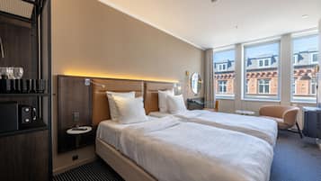 Ett hotellrum med två sängar, en minibar, ett runt bord, en stol och stora fönster med utsikt över byggnader.
