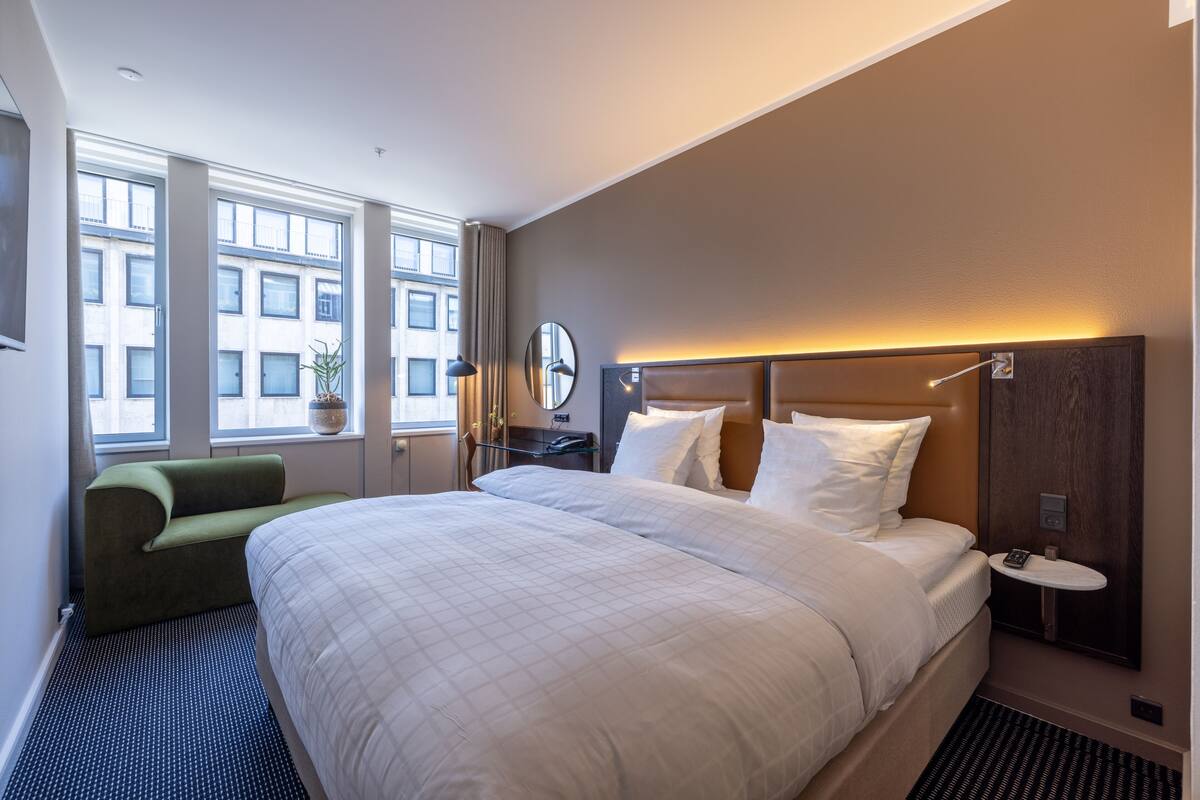 A modern hotel room with a large bed, a green armchair, a bedside table with a lamp, and a view of the city through large windows.
