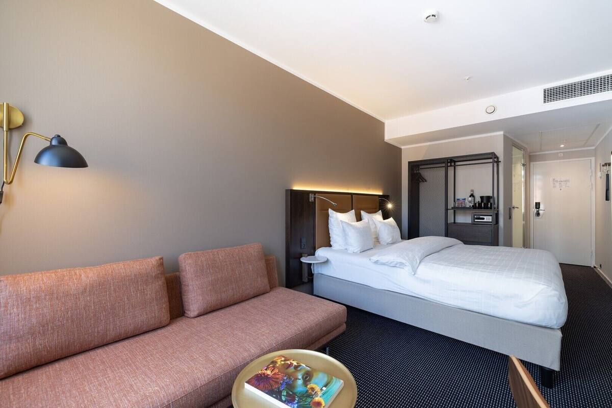 A hotel room with a bed, a sofa, a bedside table with a lamp, and a small round table with a book.