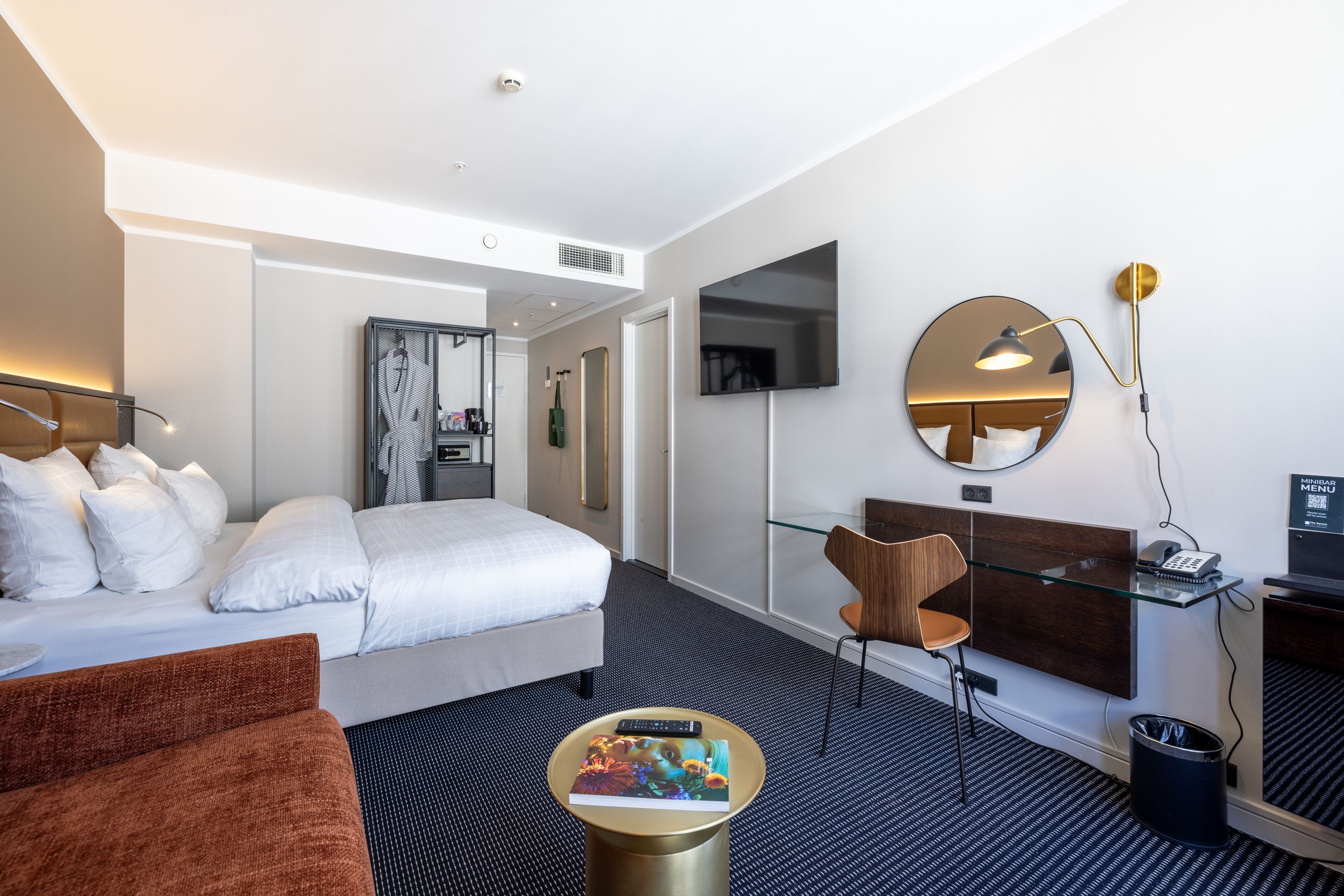 Une chambre d’hôtel moderne équipée d’un lit, d’un canapé, d’un bureau avec une chaise, d’un miroir rond et d’un téléviseur.