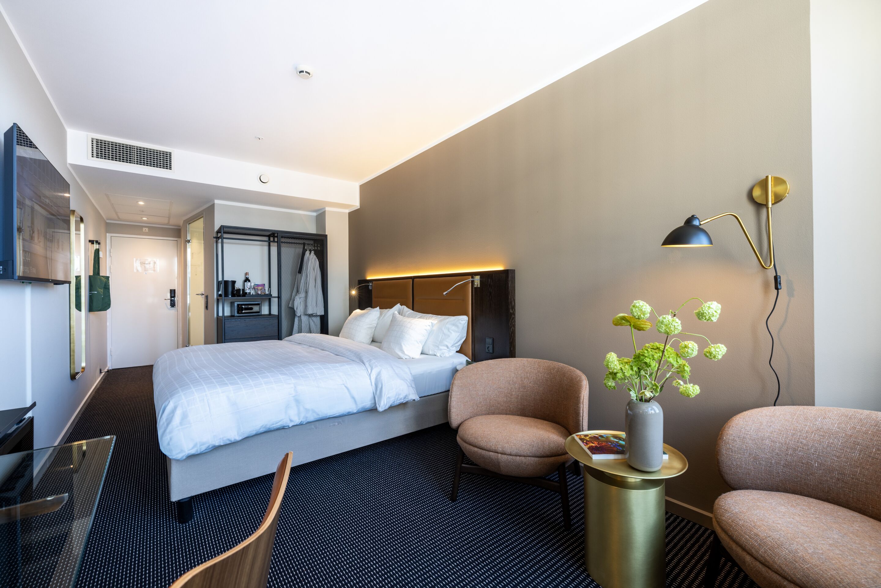 Una habitación de hotel moderna con una cama, dos sillones, una mesita de noche con un florero, un escritorio y una lámpara de escritorio.