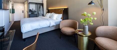 Una habitación de hotel moderna con una cama, dos sillones, una mesita de noche con un florero, un escritorio y una lámpara de escritorio.