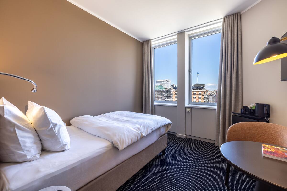 A hotel room with a bed, a desk, a chair, and a large window offering a city view.