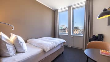 Quarto de hotel com cama, escrivaninha, cadeira e uma grande janela com vista para a cidade.