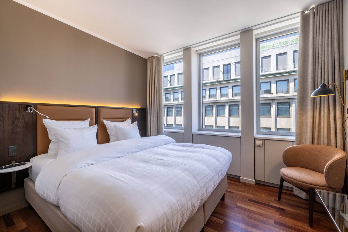 A hotel room with a large bed, a nightstand, a chair, and a window with a view of buildings.