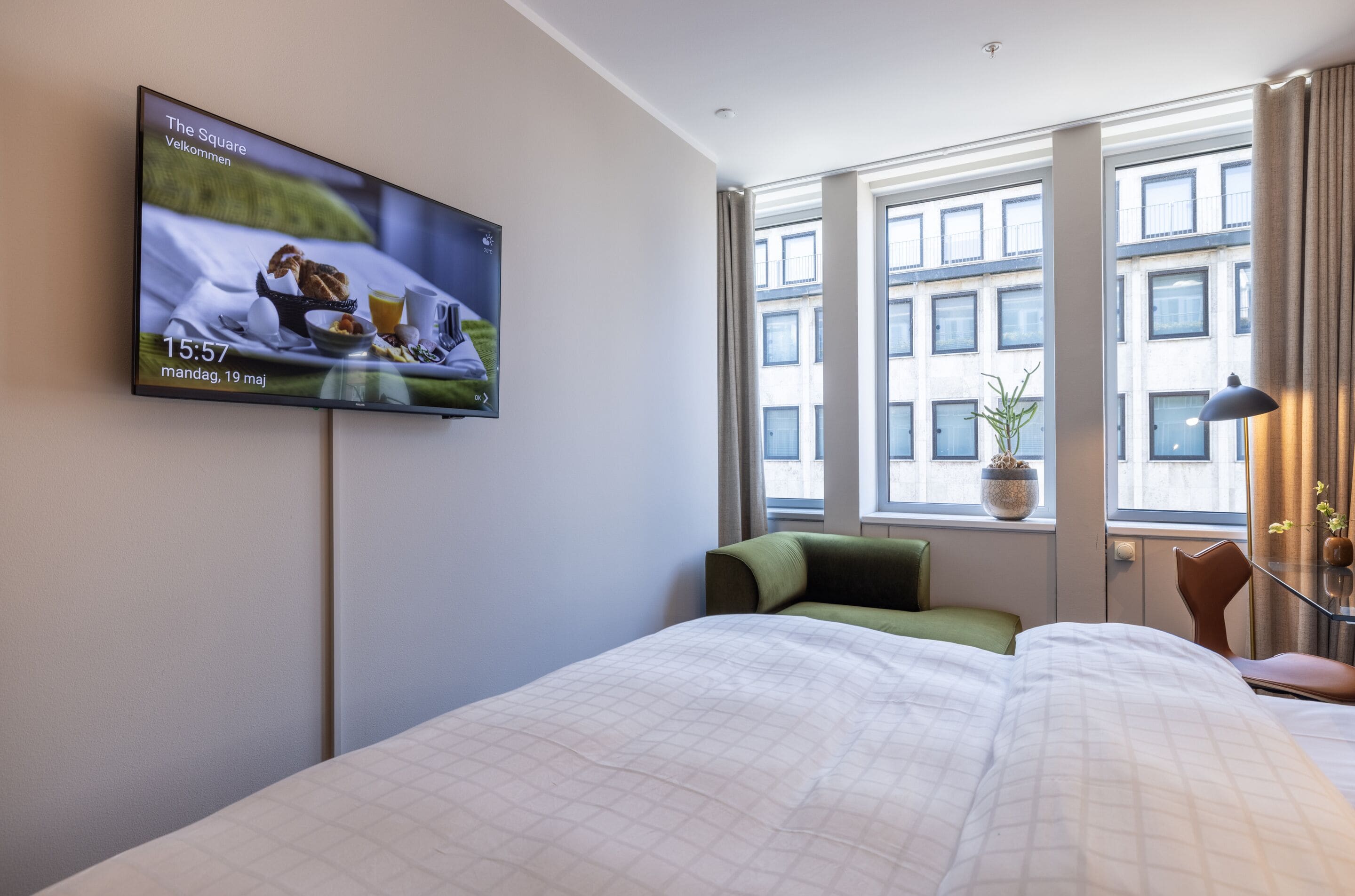 A hotel room with a bed, a TV on the wall displaying a breakfast scene, a window with a view of buildings, and a potted plant on the windowsill.