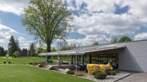 A modern building with a covered outdoor area, a large tree, and a yellow cow sculpture. - Hotel aarau-WEST (Oberentfelden)
