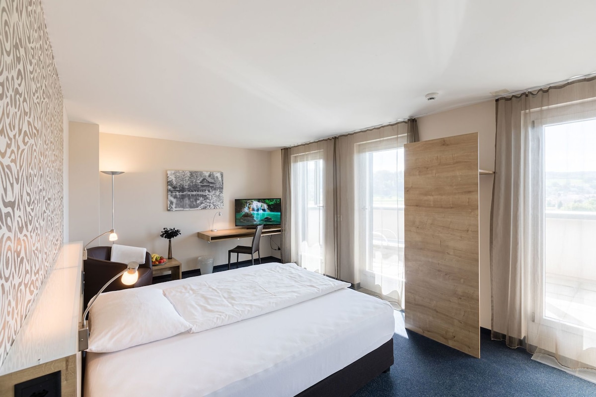 A hotel room with a bed, desk, TV, and large windows.