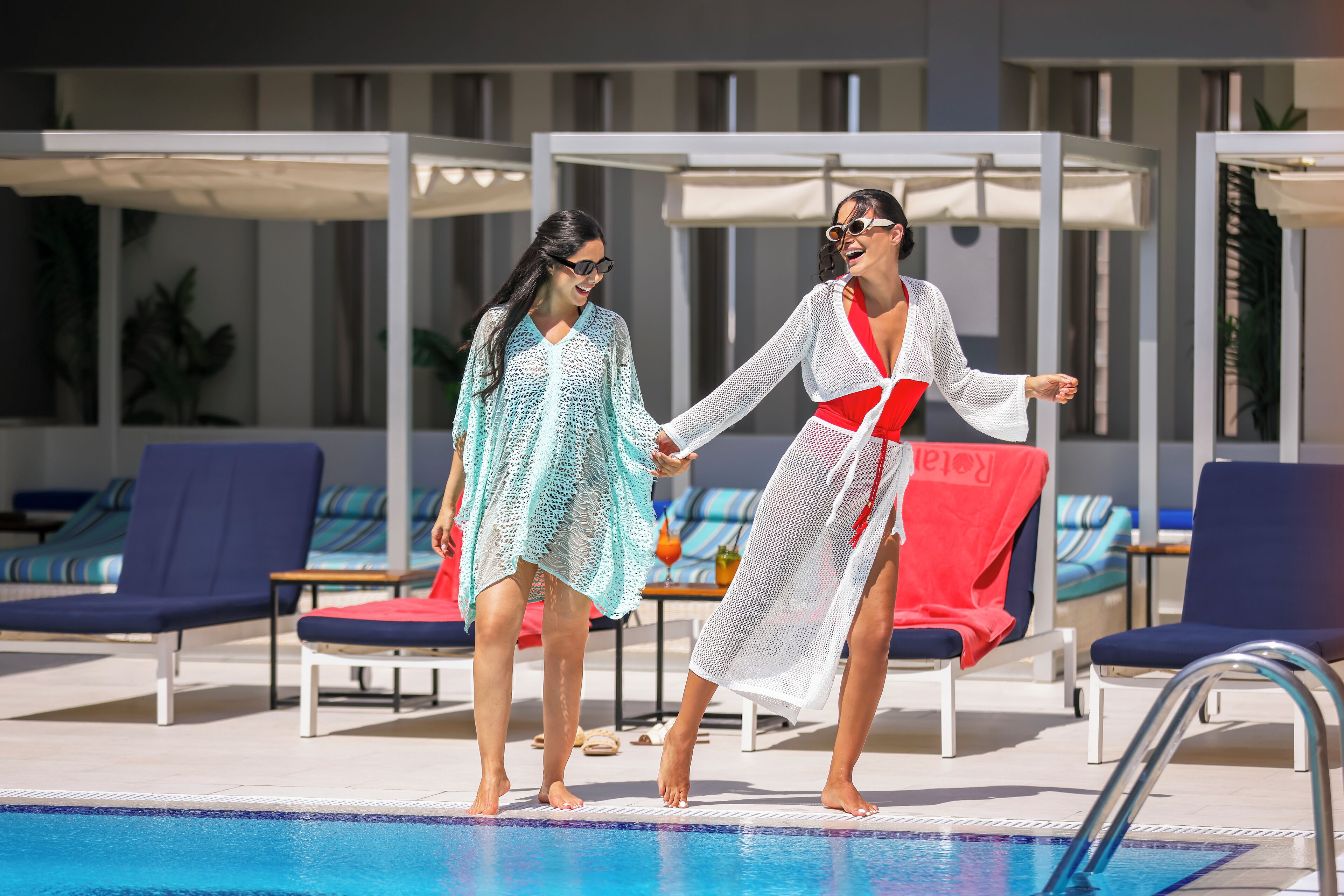 Two people holding hands by a pool with lounge chairs and umbrellas in the background.