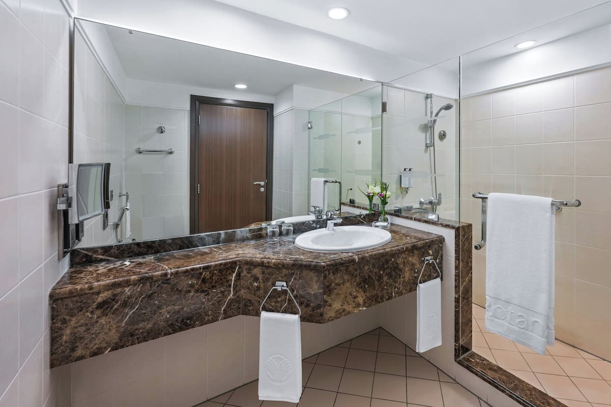 Un baño moderno con tocador de mármol, un espejo grande, cabina de ducha de vidrio y suelo de baldosas blancas.