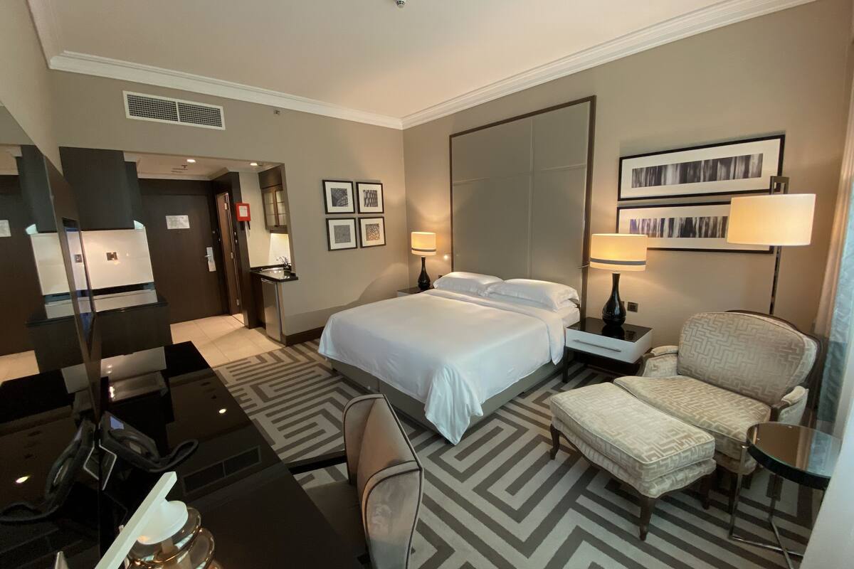 Une chambre d’hôtel avec un grand lit, deux tables de chevet avec des lampes, une chaise, une petite table et un mini-bar.