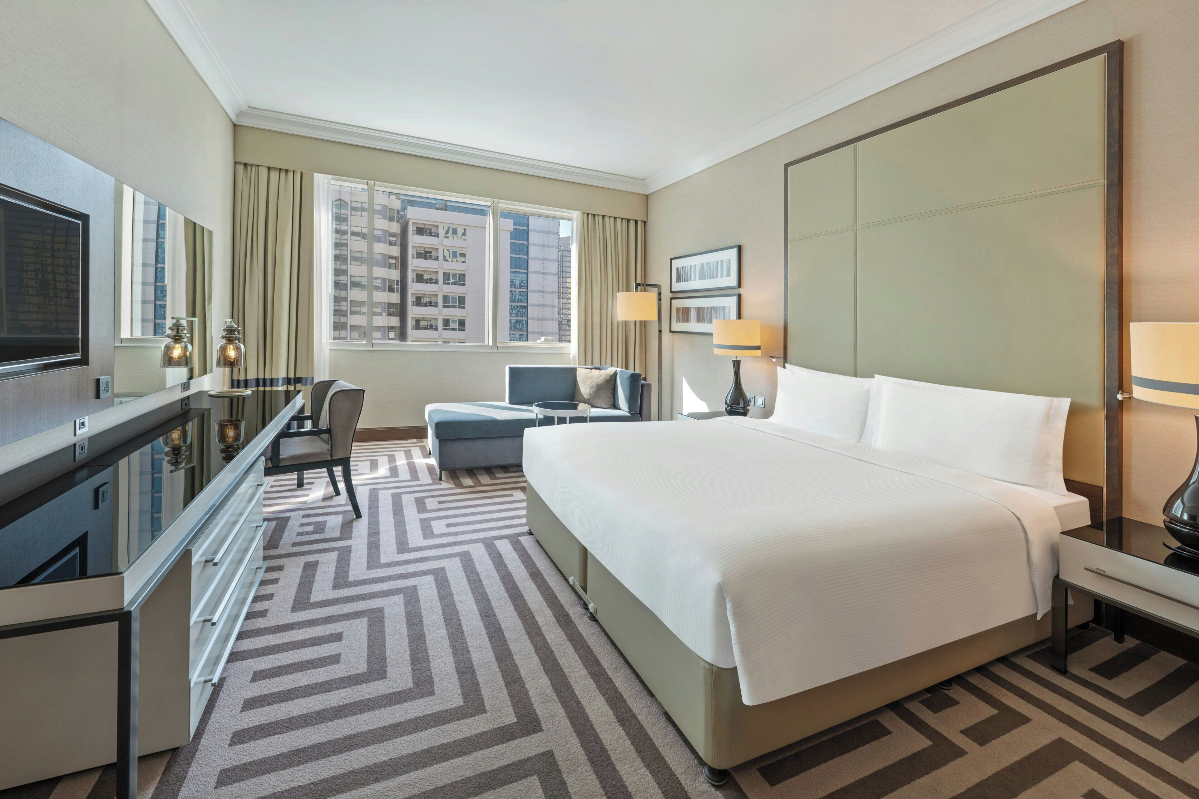 A hotel room with a large bed, a desk with a chair, a TV, and a view of the city.