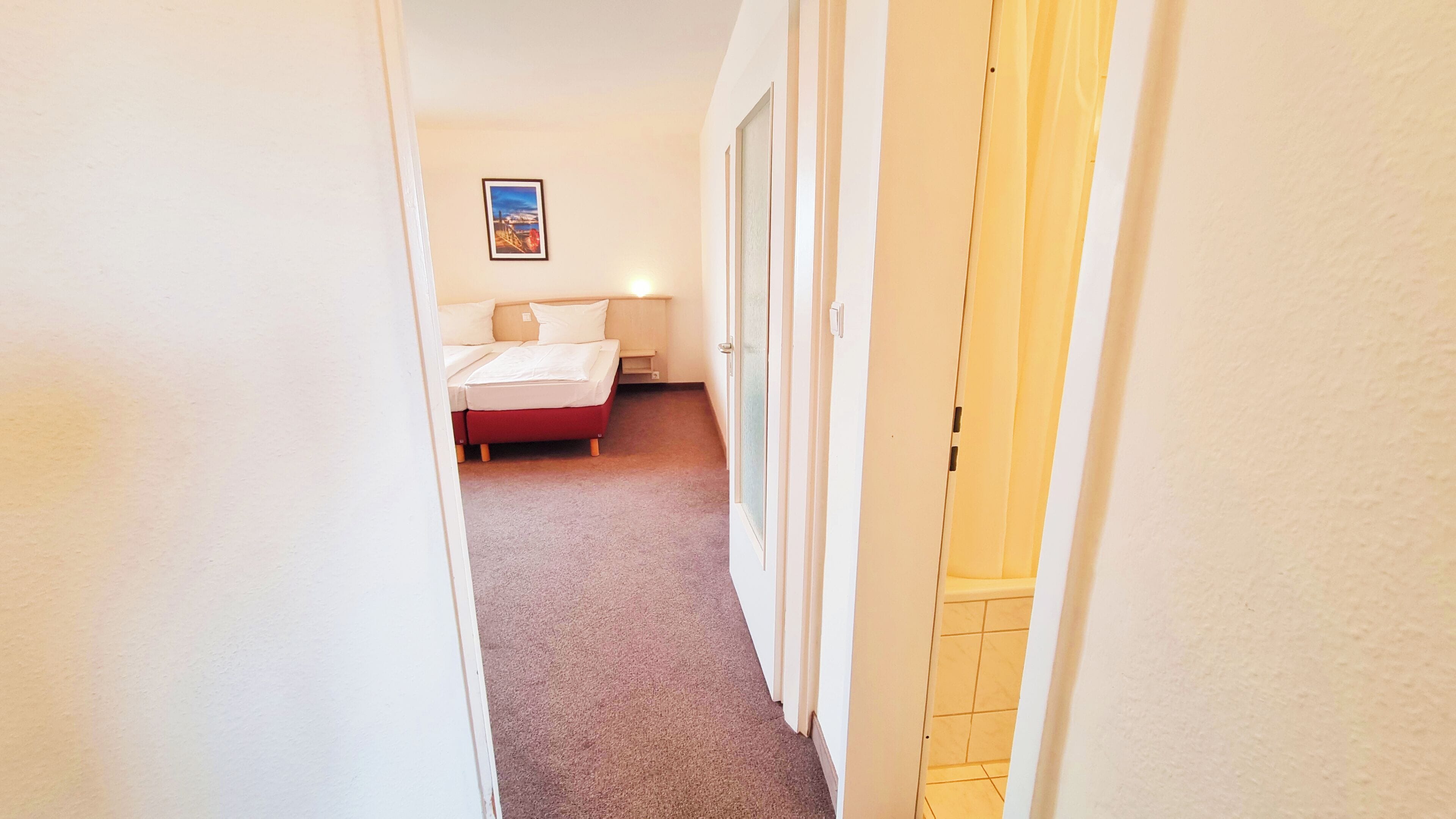 A narrow hallway leading to a bedroom with a bed, a nightstand, and a framed picture on the wall.