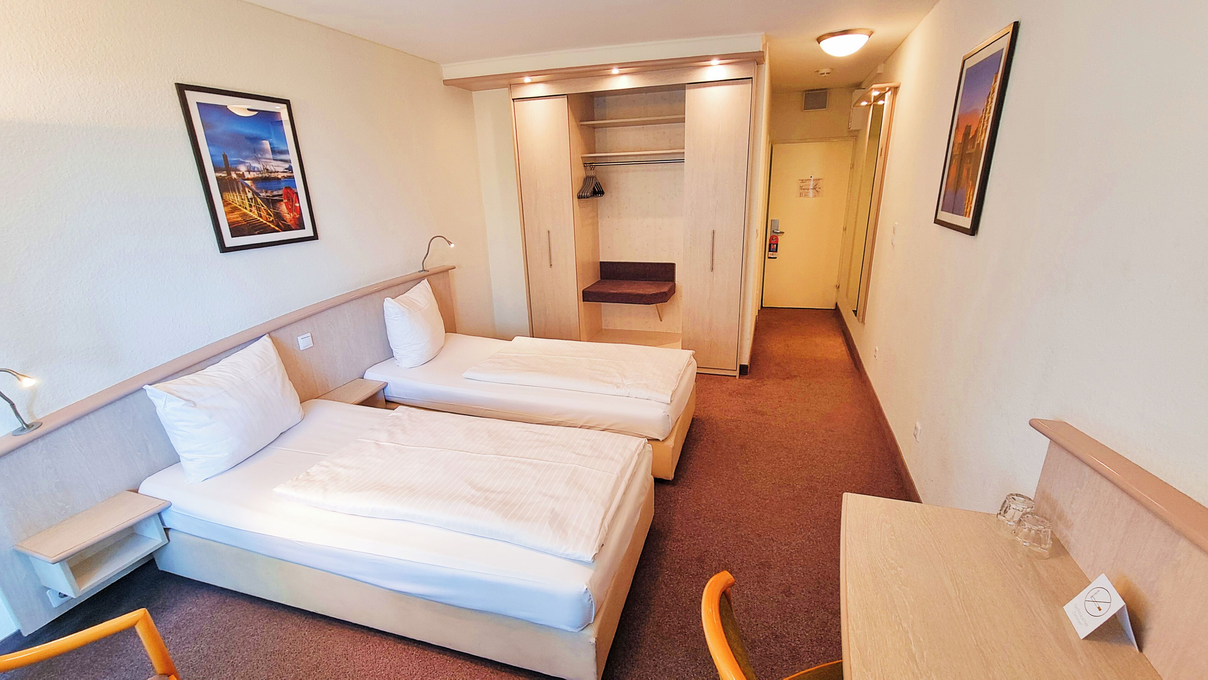 A hotel room with two beds, a small table, a chair, a wardrobe, and a corridor leading to another room.