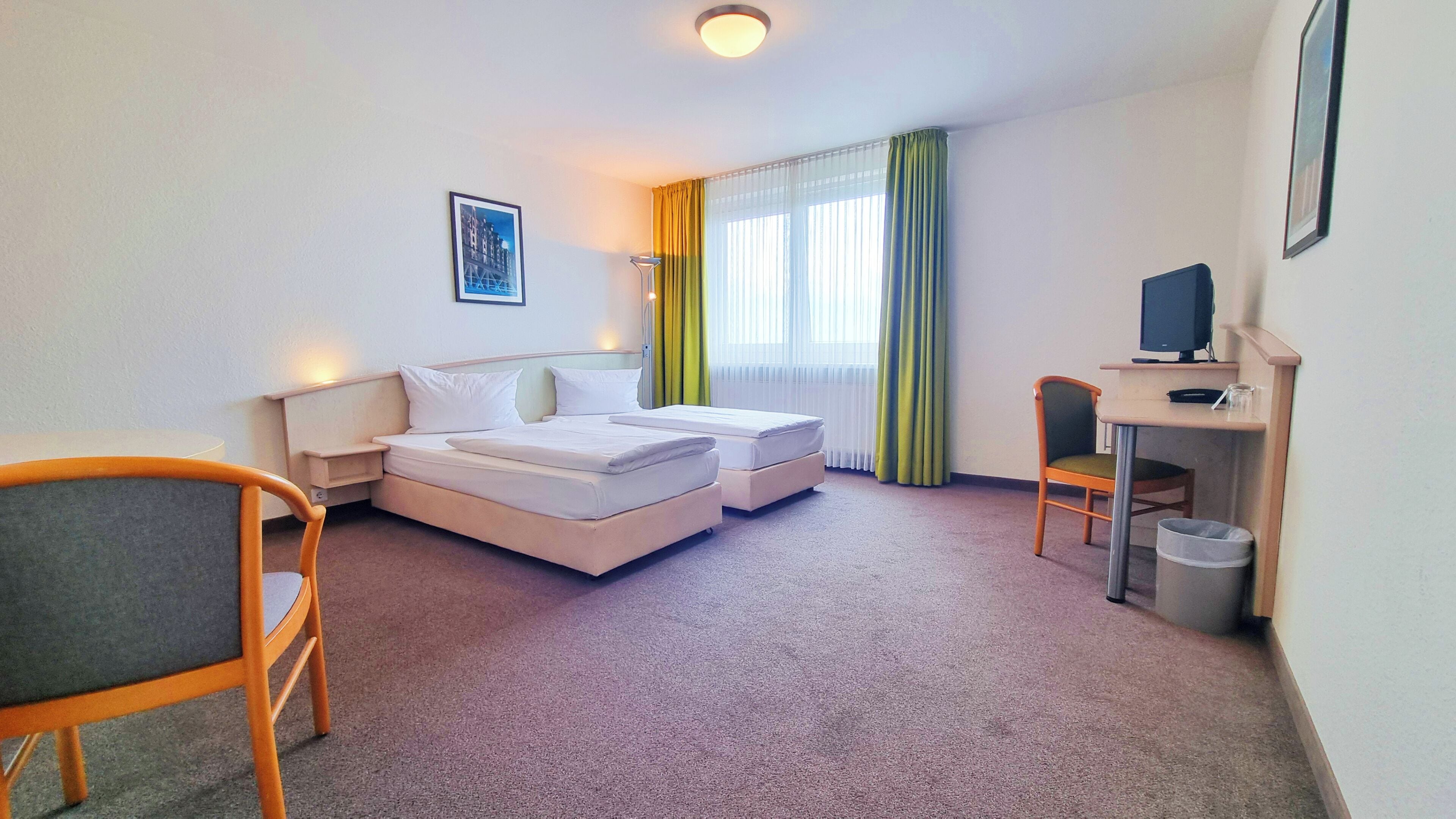 A hotel room with a bed, a desk with a computer, a chair, and a small trash bin.