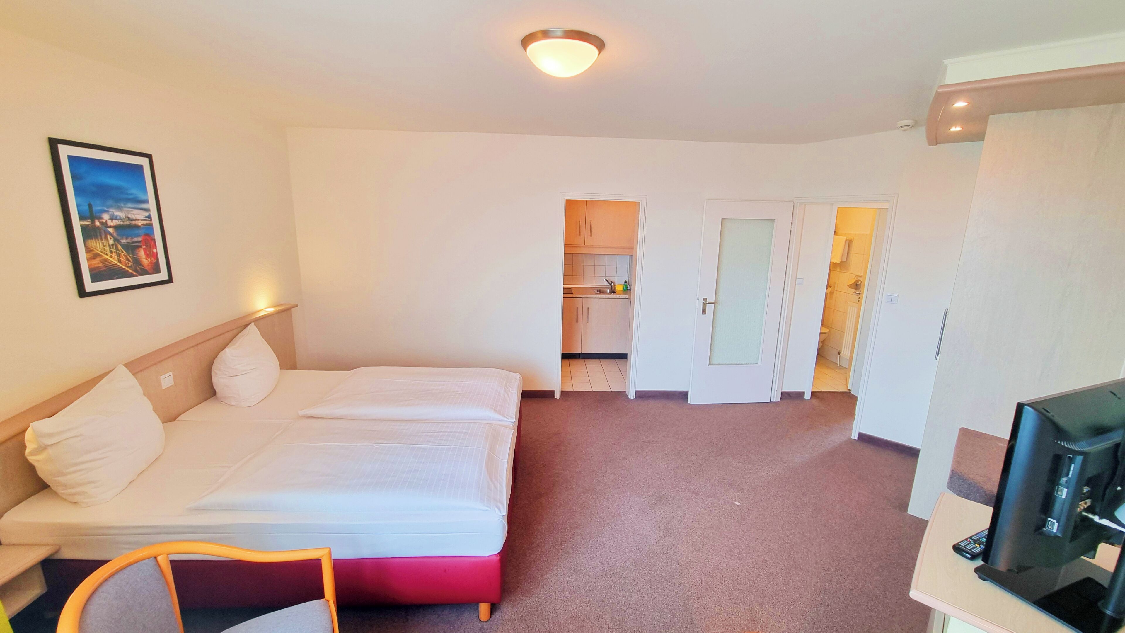 A hotel room with two beds, a TV, a desk, a chair, a picture on the wall, and a kitchen area visible through an open door.