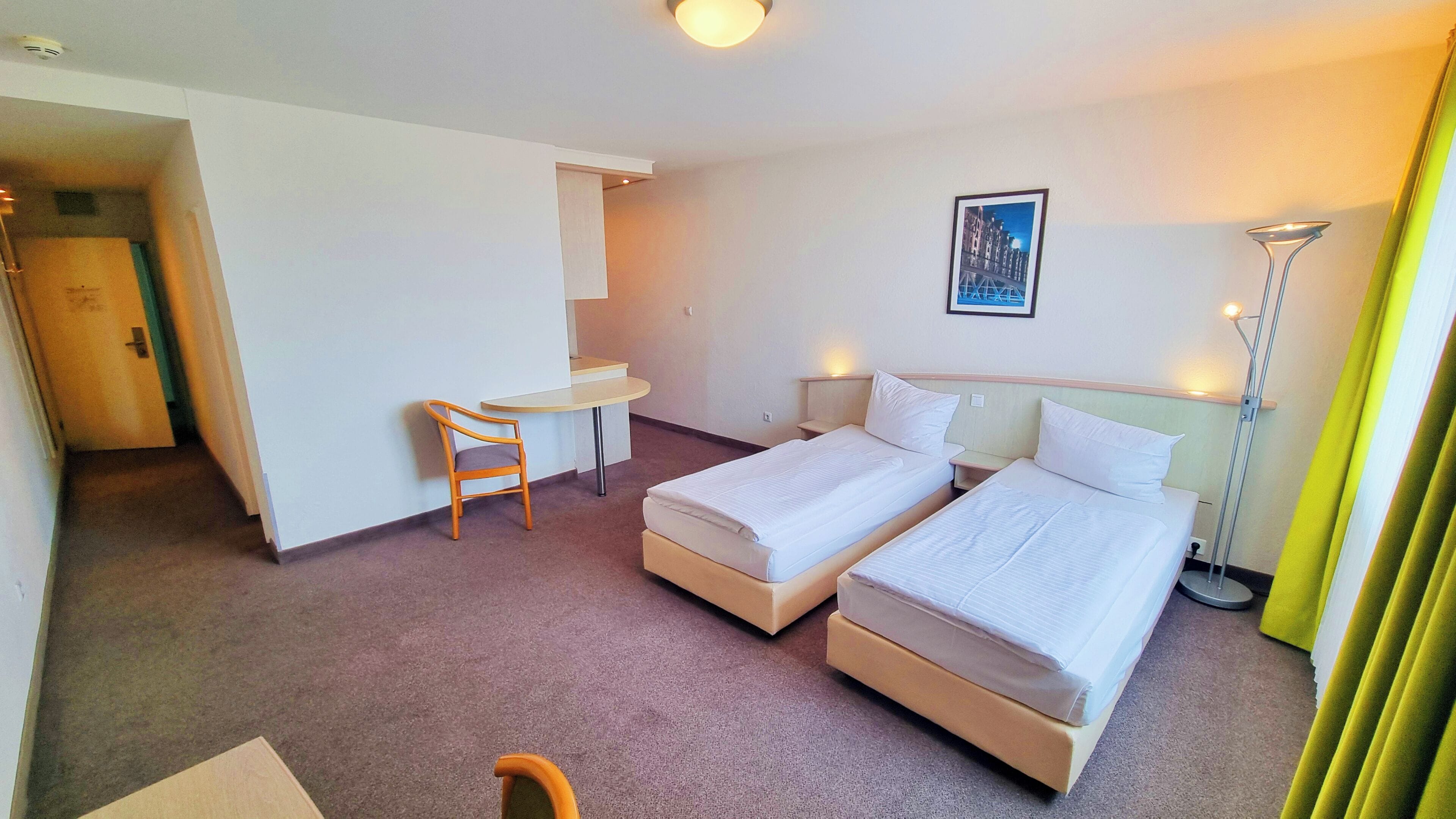 Ein Hotelzimmer mit zwei Einzelbetten, einem kleinen Schreibtisch, einem Stuhl, einem Nachttisch, einer Lampe und einem gerahmten Bild an der Wand.