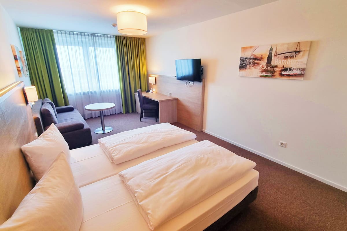 A hotel room with a large bed, a desk with a chair, a small table, a TV mounted on the wall, and a painting of a cityscape.