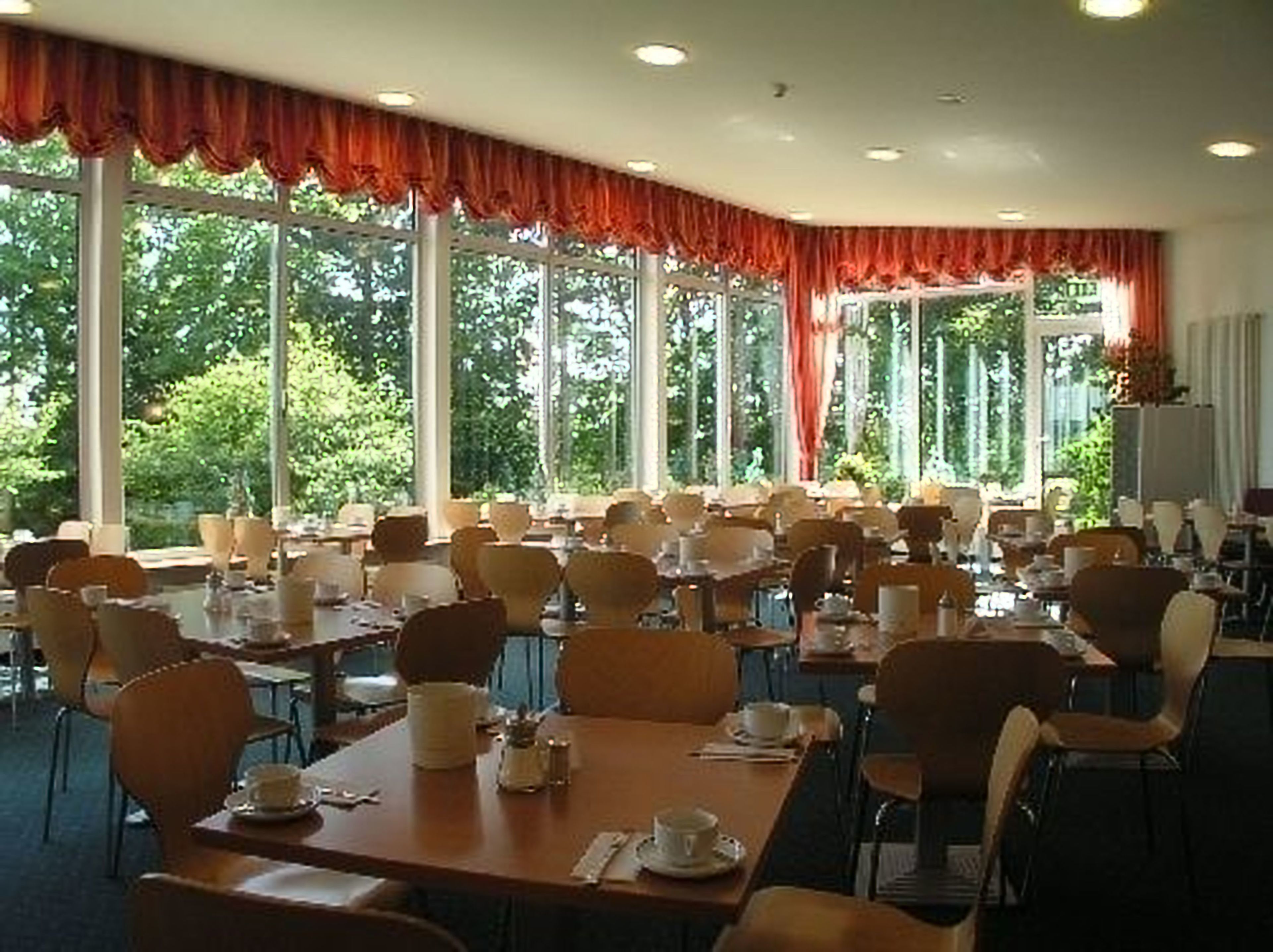 A dining area with tables and chairs, large windows with curtains, and a view of greenery outside.