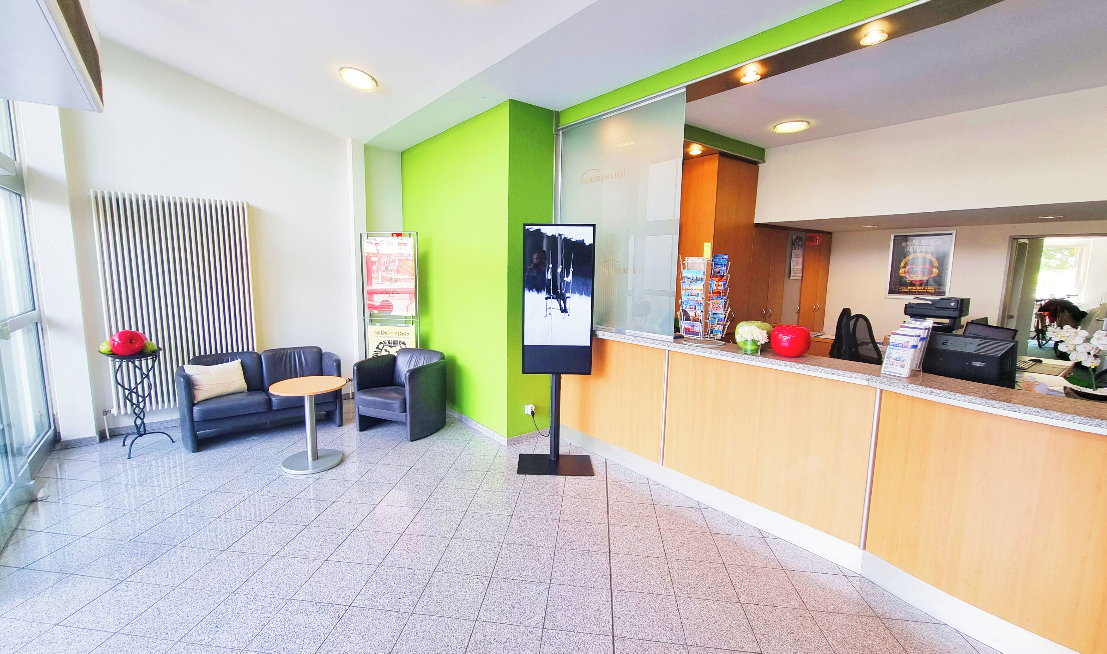 Un área de recepción con una pared de acento verde, un mostrador de recepción con una computadora e impresora, un rincón de espera con sofá y sillones, y un tablón de anuncios con un mapa.