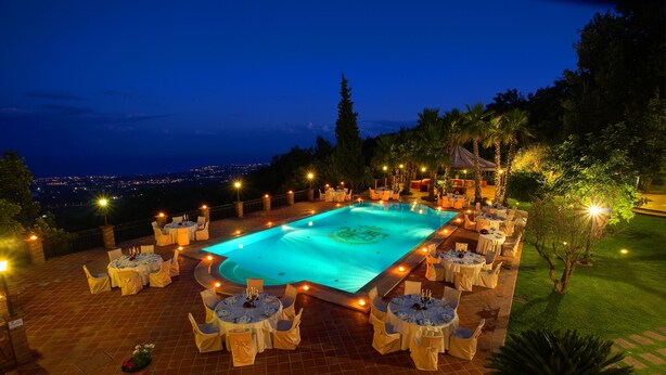 An outdoor pool area with tables and chairs arranged around it, surrounded by trees and a view of the city in the background. - Airone Wellness Hotel (Zafferana Etnea)