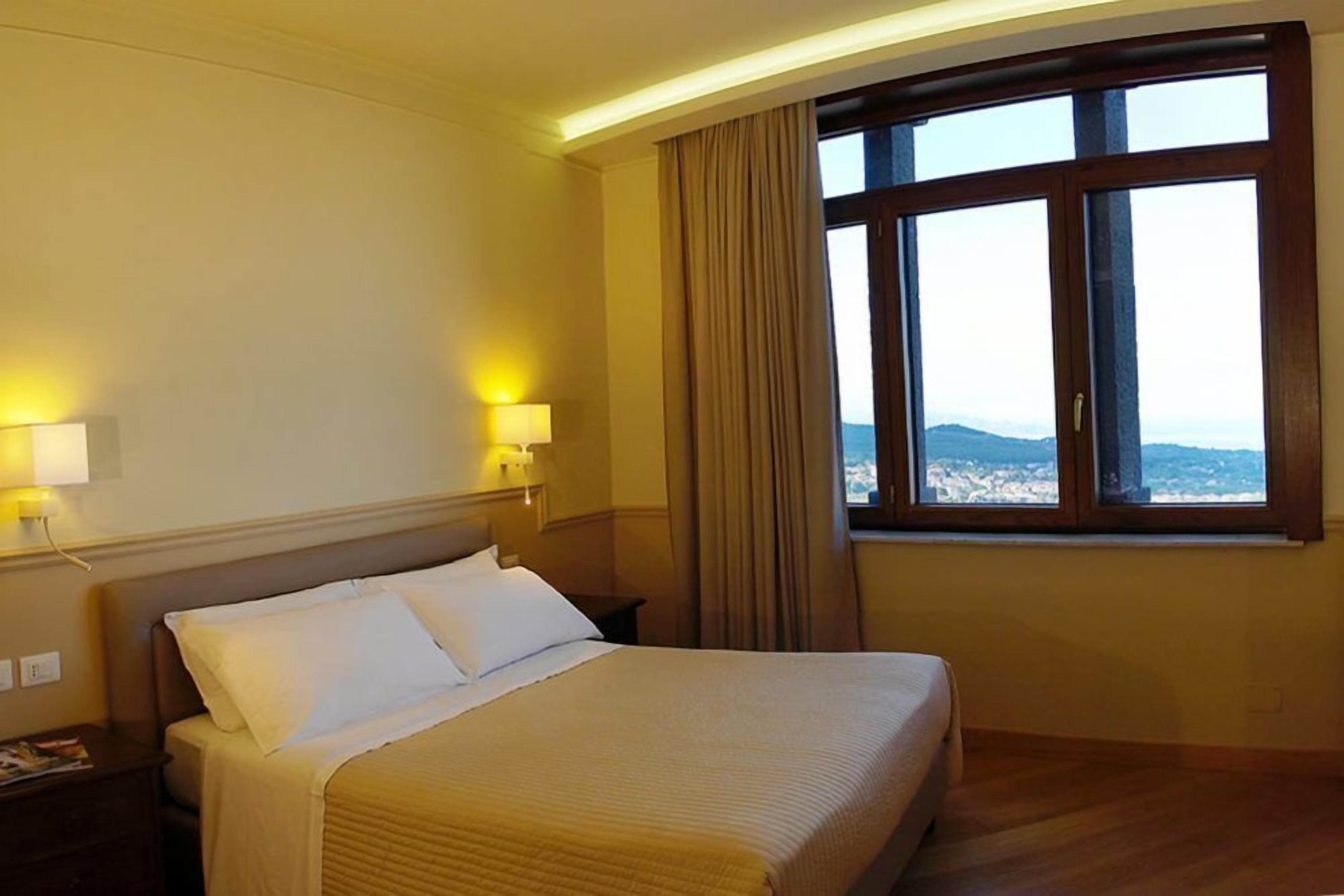 Hotelkamer met een groot bed, twee bedlampen, een raam met gordijnen en uitzicht op de bergen.