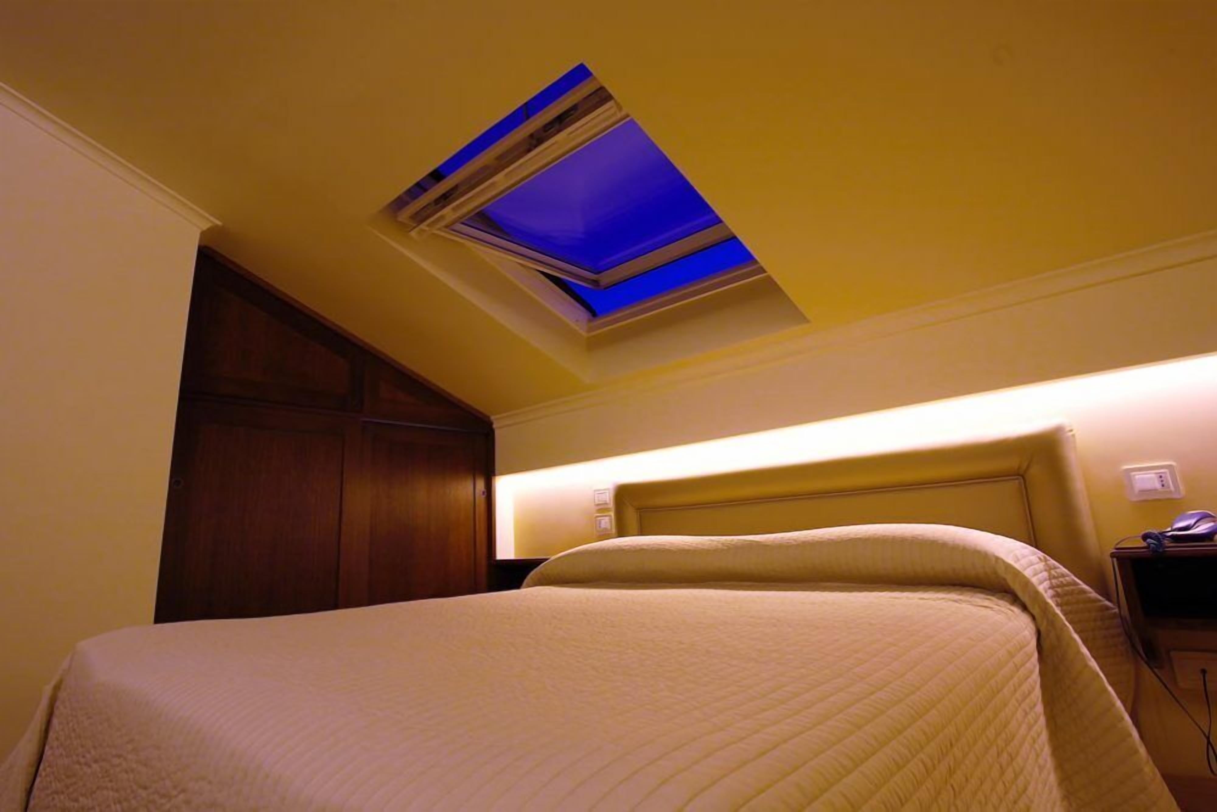 Habitación de hotel con cama, mesita de noche, armario de madera y tragaluz con iluminación azul.