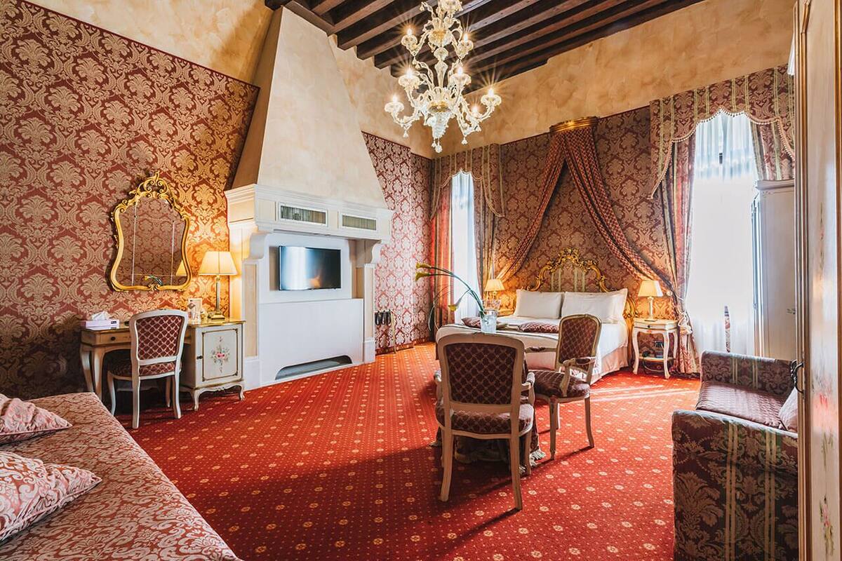 Habitación de hotel con alfombra roja, una cama, escritorio, silla, televisor y candelabro.