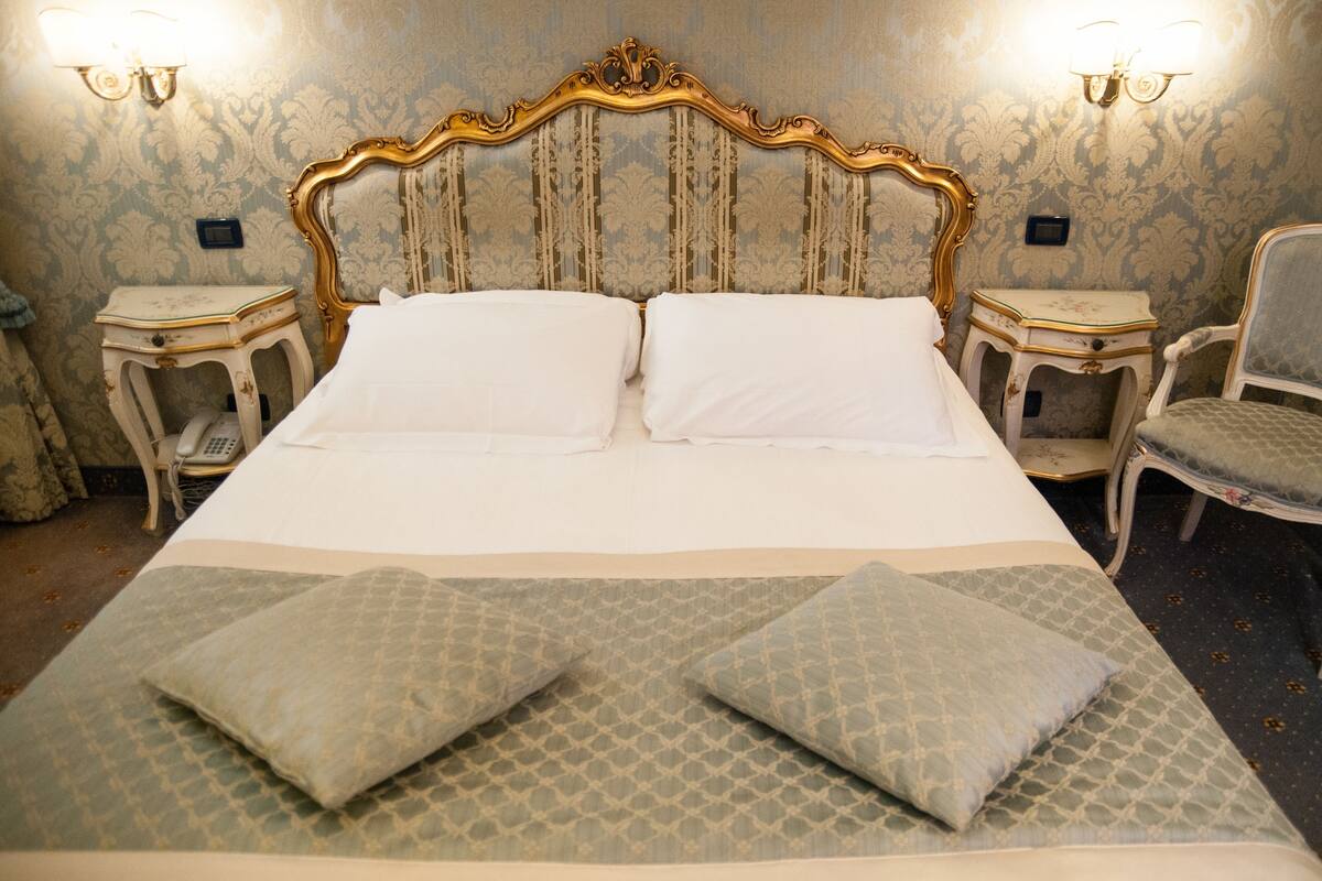 Una cama grande con cabecera dorada, dos mesitas de noche y una silla.