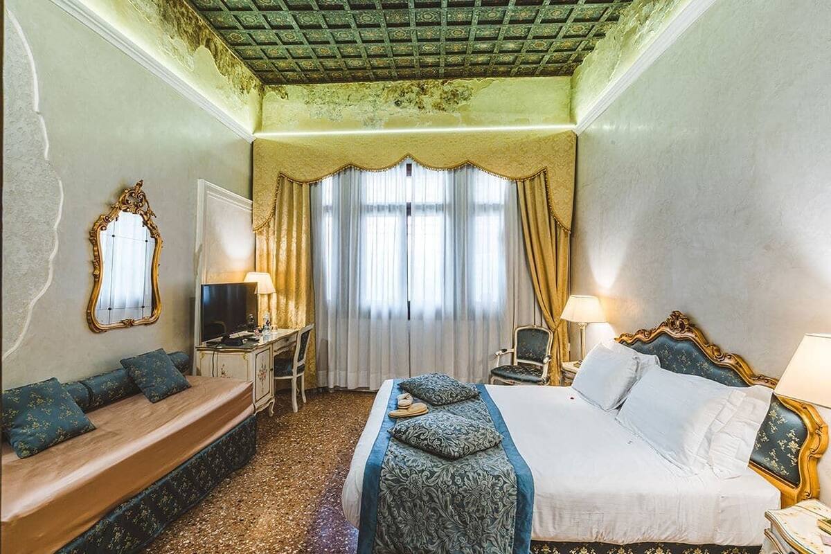 Habitación de hotel con cama, un banco, escritorio con televisor y un amplio ventanal con cortinas.